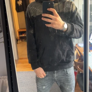 Svart hoodie med silvervingar från Marcelo Burlon - Svart hoodie från Marcelo Burlon med ikoniskt silverfärgat vingtryck över axlar och bröst. Tröjan har huva med snörning, känguruficka och ribbade muddar. Perfekt statement-plagg för dig som vill sticka ut.