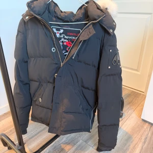 Svart dunjacka Moose Knuckles - Svart dunjacka från Moose Knuckles med pösig passform, avtagbar huva med pälsdetalj och ikonisk logga på ärmen. Jackan har dragkedja, knappar och flera fickor. Perfekt för kalla dagar och riktigt snygg streetstil.