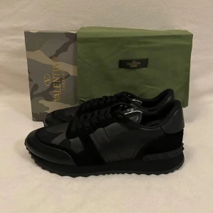 Valentino Garavani Rockrunner sneakers - Snygga svarta Valentino Garavani Rockrunner sneakers med diskret camouflage-mönster. Skorna har en mix av skinn och mocka, klassisk snörning och en chunky sula med coola nitar baktill. Perfekta för dig som vill ha en exklusiv och stilren look.