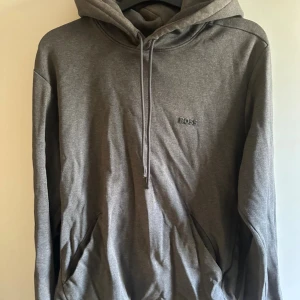 Grå hoodie från BOSS med huva - Säljer en grå hoodie från BOSS med broderad logga på bröstet. Tröjan har huva med snörning och en stor magficka. Materialet är mjukt och bekvämt, perfekt för chill dagar. Långärmad och loose passform för en avslappnad stil.