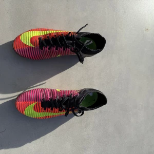 Nike Mercurial randiga fotbollsskor - Säljer ett par Nike Mercurial fotbollsskor med coolt randigt mönster i svart, rött och rosa med neongula detaljer och snörning. Skorna har högre strumpliknande skaft och plös, samt dobbar för gräsplan. Perfekta för dig som vill sticka ut på planen.