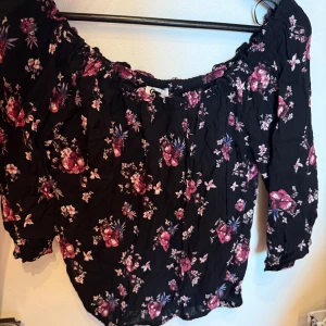 Svart blommig offshoulder blus - Säljer en svart blus med offshoulder och långa ärmar. Blusen har ett blommigt mönster i rosa och lila toner, samt en lätt skrynklig struktur. Perfekt för dig som gillar romantiska vibes och vill ha något som sticker ut.