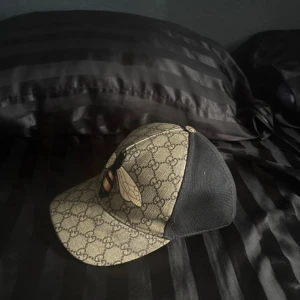 Gucci keps med bi och GG-mönster - Snygg keps från Gucci med klassiskt GG-mönster i beige och svart. Framsidan pryds av en stor broderad bi och baksidan är i svart mesh för extra ventilation. Justerbar rem med gröna och röda detaljer. Perfekt accessoar för att sticka ut.