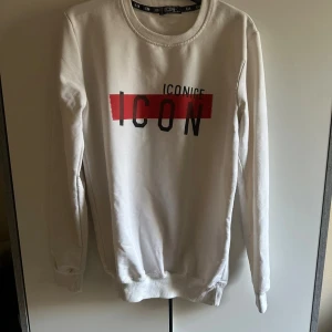 Icon tröja - Säljer en vit sweatshirt från ICONIC med tryck i svart och rött över bröstet. Tröjan har rund hals, långa ärmar och ribbade muddar vid ärmslut och nederkant. Perfekt för dig som gillar streetstyle och enkel design.