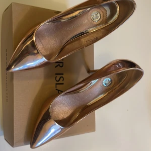 Metallic roséguld pumps River Island - Snygga pumps i metallic roséguld från River Island. Skorna har spetsig tå, hög klack och en glansig finish som verkligen sticker ut. Perfekta för dig som vill ha ett par statement-heels med elegant vibe. Tillverkade i syntetmaterial.