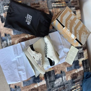 Golden Goose Superstar vita sneakers - Golden Goose Superstar sneakers i vitt skinn med grå mockastjärna på sidan, svarta detaljer vid hälen och grå snörning. Klassisk låg modell med platt sula och rund tå. Snygg och stilren design som passar till det mesta.