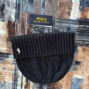 Svart kabelstickad mössa Polo Ralph Lauren - Svart kabelstickad mössa från Polo Ralph Lauren med bred ribbad uppvikning och klassisk broderad logga i vitt framtill. Tillverkad i mjuk ullblandning med 26% återvunnen nylon. One size och perfekt för kyliga dagar.