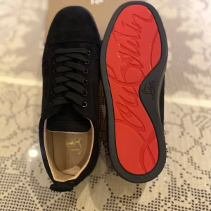 Christian Louboutin svarta sneakers mocka - Snygga svarta sneakers från Christian Louboutin i mjuk mocka med klassisk röd sula och diskret logga. Skorna har rund tå, svarta snören och stilren design som sticker ut med den ikoniska röda undersidan. Perfekta för dig som gillar lyxiga detaljer och streetstyle.
