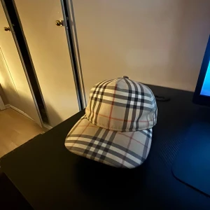 Burberry rutig keps med logga - Säljer en klassisk keps från Burberry med det ikoniska rutiga mönstret i beige, svart, vitt och rött. Kepsen har böjd skärm och justerbar baksida med svart patch där det står 'Burberry London England'. Perfekt accessoar för en trendig look.