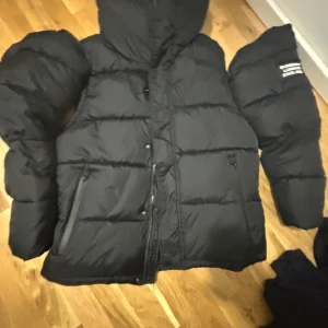 Svart pufferjacka från Burberry - Säljer en svart pufferjacka från Burberry med stor huva och tydlig logga på ärmen. Jackan har dragkedja och knappar framtill samt två sidofickor med dragkedja. Perfekt för kalla dagar och riktigt snygg streetstil.