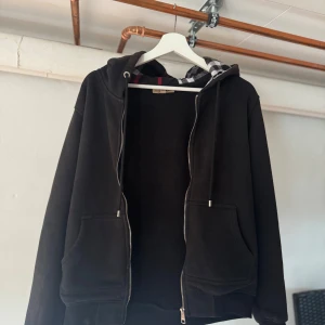 Svart hoodie med dragkedja och rutigt foder - Svart hoodie från Burberry med dragkedja framtill, fickor och huva med klassiskt rutigt foder. Tröjan har långa ärmar och snörning i huvan. Perfekt för dig som gillar stilren streetwear med en touch av lyx.