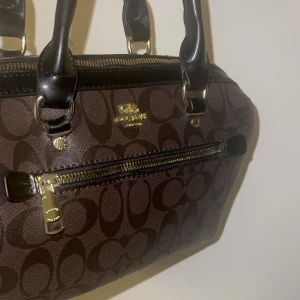 Coach medium väska  - Snygg brun handväska från Coach med klassiskt monogrammönster och svarta handtag. Väskan har guldfärgade detaljer, dragkedja upptill och ett extra fack med dragkedja framtill. 