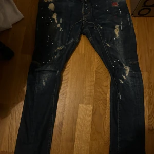 Mörkblå DSQ2  jeans med slitningar och färgstänk - Säljer ett par mörkblå DSQ2 jeans med coola slitningar och vita färgstänk för en edgy look. Jeansen har smal passform, klassiska bakfickor och kontrastsömmar. Perfekta för dig som gillar streetstyle och vill sticka ut lite extra.