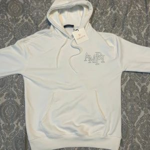 Vit hoodie från Amiri med broderi - Säljer en stilren vit hoodie från Amiri med stort broderat AMIRI-logo på både bröstet och ryggen. Tröjan har huva med snörning och en klassisk känguruficka framtill. Perfekt för dig som gillar exklusiva streetwear-plagg.