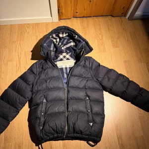 Svart pufferjacka från Burberry - Snygg svart pufferjacka från Burberry med klassiskt rutigt foder i blått och vitt. Jackan har huva, dragkedja framtill och två fickor med tryckknappar. Perfekt för kalla dagar och har en riktigt clean look.