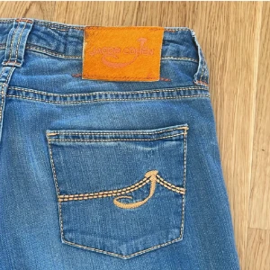 Blå jeans från Jacob Cohën,  W28 - Snygga blå jeans från Jacob Cohën med klassisk femficksdesign och coola kontrastsömmar. Jeansen har ett broderat märke på bakfickan och en orange patch i midjan. Tillverkade i mjukt denimtyg för en skön känsla.