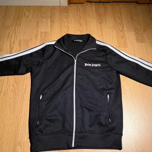 Svart track jacket från Palm Angels - Svart track jacket från Palm Angels med vita ränder längs ärmarna och vit dragkedja. Jackan har hög krage, två fickor med dragkedja och logga på bröstet. Perfekt för en sportig och trendig look.