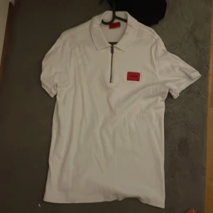 Vit HUGO t-shirt med dragkedja - Snygg vit t-shirt från HUGO med klassisk krage och halv dragkedja framtill. Röd logotyp-patch på bröstet ger en clean men ändå sportig vibe. Perfekt till jeans eller shorts för en fräsch look.