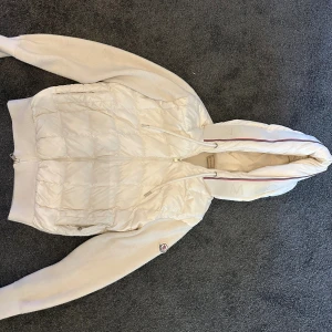 Vit Moncler Cardigan med huva - Säljer en snygg vit Cardigan från Moncler med huva och ribbstickade ärmar. Jackan har quiltad front, dragkedja och diskret Moncler-logga på ärmen. Perfekt för dig som vill ha en clean och exklusiv look med street-vibe. (Priset kan diskuteras)