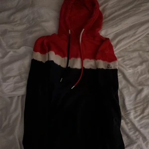 Moncler hoodie i rött, vitt och svart - Säljer en snygg hoodie från Moncler med färgblock i rött, vitt och svart. Tröjan har huva med snörning och Moncler-logga på bröstet. Perfekt för dig som vill ha en streetig och trendig look.