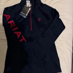 Ariat fleece strl XS - Tek team 1/2 zip swtshirt navy heather.  Tunn funktionsfleece från Ariat. Material som andas och transporterar bort väta från kroppen. Halv ståkrage och blixtlås fram gör tröjan lätt att dra på och av. Ergonomiska hål för tummarna. Broderad logo på ärmen. Strl xs.