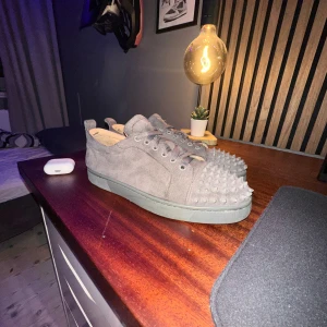 Grå mocka sneakers med nitar - Säljer ett par gråa sneakers i mocka med coola nitar över tån och platt sula. Skorna har snörning och en unik röd undersida som sticker ut. Perfekt för dig som vill ha något edgy och annorlunda i din skogarderob. Skriv för mer bilder vill byta mot ett par i st 39