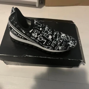 Svart-vita slip-on sneakers från DKNY - Säljer ett par svarta och vita slip-on sneakers från DKNY i modellen Aliza. Skorna har ett grafiskt mönster med grov, räfflad sula. Storlek 38. Skorna är nya, men lådan är smått skadad.