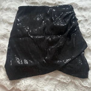 Svart paljettkjol från Pimkie - Snygg svart kortkjol från Pimkie med glittriga paljetter över hela ytan. Kjolen har omlott-design framtill och är perfekt för dig som vill sticka ut med lite extra bling. Materialet är mjukt och följsamt, vilket gör den bekväm att bära. 