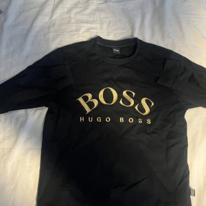 Svart tröja från Hugo Boss med guldtryck - Svart långärmad tröja från Hugo Boss med stort guldigt BOSS-tryck på bröstet. Klassisk rund halsringning och ribbade muddar. Perfekt för dig som gillar stilrena och lyxiga plagg med tydlig logga.