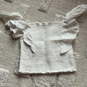 Vit volangblus - Supersöt vit blus med smockad kropp och breda volangärmar. Toppen har en fyrkantig halsringning och små volanger längst ner. Materialet känns som bomull och är lätt och luftigt, perfekt för varma dagar. 