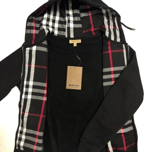 Burberry kofta svart  - Svart kofta från Burberry 10/10 kvalité med klassiskt rutig insida i svart, vitt och rött. Koftan har huva med dragsko, dragkedja framtill och diskret broderad logga på bröstet. Perfekt för de kommande kyliga dagar. Storleken är M. Skickas inom 24timmar! 