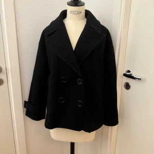 Svart skepparkavaj från Design by Si - XS/S Stilren svart skepparkavaj från Design by Si med dubbelknäppning och bred krage. Jackan har långa ärmar med knappdetalj vid mudden och en klassisk, boxig passform. 