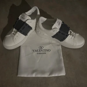 Valentino Garavani Rockstud sneakers - Säljer ett par vita Valentino Garavani Rockstud sneakers med mörkblå band och ikoniska nitar över vristen. Skorna har snörning, platt sula och rund tå. Tillverkade i skinn med exklusiv känsla och kommer med original dustbag. Hör av vid minsta fundering 💫