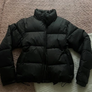 Svart pufferjacka från Everest - Svart pufferjacka från Everest med hög krage och dragkedja framtill. Jackan har en quiltad design och justerbar nederkant med snörning. Perfekt för kalla dagar och har en clean, stilren look som funkar till allt.