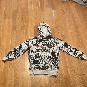 Vit hoodie med svart graffiti från Woxo - Säljer en vit hoodie från Woxo 720° med svart graffiti-inspirerat tryck över hela tröjan och en röd linje på bröstet. Tröjan har huva, långa ärmar och ribbade muddar. Perfekt för dig som gillar streetstyle och coola prints.
