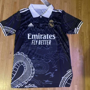 Real Madrid x Adidas svart fotbollströja - Snygg svart Real Madrid fotbollströja från Adidas med vit krage och detaljer. Tröjan har ett stort drakmotiv över hela framsidan och baksidan, samt klubbmärke och sponsortryck på bröstet. Tillverkad i lätt och ventilerande material, perfekt för match eller träning.