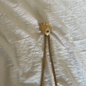 Vintage guldigt halsband med grön sten - Unikt halsband i guldton med en dekorativ medaljongformad hänge som har ett fjäderliknande mönster. Hänget är prytt med en grön sten och små klara stenar. Kedjan är kraftig och matchar hängets guldiga färg. Perfekt för dig som gillar statement-smycken.