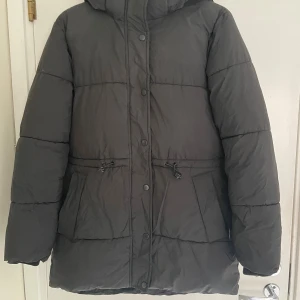 Svart pufferjacka från new yorker  - Svart pufferjacka från Amisu med huva, stora fickor och snörning i midjan. Jackan har quiltad design och stängs med både dragkedja och knappar. Perfekt för kalla dagar och har en loose passform som ger skön streetkänsla. Sitter som S. Har ett litet hål (se bild 4) men de syns knappt