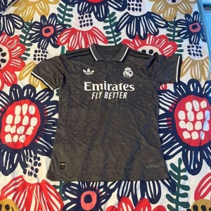 Real Madrid x Adidas grå fotbollströja - Snygg grå Real Madrid fotbollströja från Adidas med klassisk krage och vita detaljer. Tröjan har korta ärmar, broderat klubbmärke och Adidas-logga samt 'Emirates Fly Better' tryckt på bröstet. Tillverkad i lätt och ventilerande material med diskret mönster i tyget. Det är spelar version så det är då slim fit