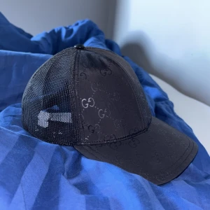 Svart meshkeps från Gucci - Snygg svart keps från Gucci med klassiskt GG-mönster över hela framsidan. Kepsen har meshpaneler på sidorna och bak, samt justerbar snapback. Framsidan är i blankt tyg och baksidan har en läderdetalj med Gucci-logga. Perfekt för en trendig streetstyle-look. Storlek L men liten i form perfekt för s-m