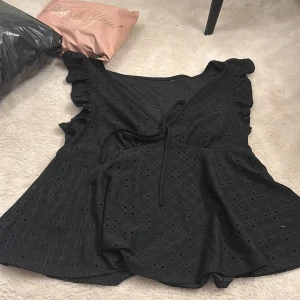 Svart blus med spets och snörning - Säljer en svart blus med v-ringning, spetsdetaljer och broderat hålmönster. Blusen har korta volangärmar och en söt snörning framtill som ger extra stilpoäng. Står inte vart den är köpt💕