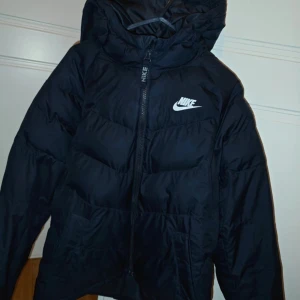 Svart pufferjacka från Nike med huva - Svart pufferjacka från Nike med stor huva och vit logga på bröstet. Jackan har dragkedja framtill med Nike-tryck och är quiltad för extra värme. Köpt förra vintern så är inte mycket använd. Den är hel och ren. Köpt för 1399:-  står att den är i storlek 150-158, passar även mig som har xs.