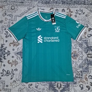 Liverpool Adidas fotbollströja Isak XL - Liverpool tredjeställ 2025/26 fotbollströja i blågrön färg från Adidas med vita detaljer och broderat klubbmärke. Nummer 9 och namnet Isak på ryggen. Klassiska Adidas-ränder på axlarna och vit-svart ribbad krage. Tillverkad i lätt och ventilerande material.