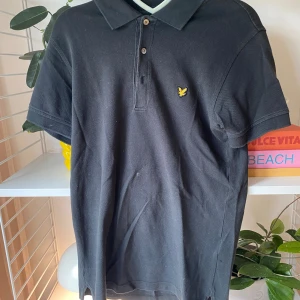 Svart piké från Lyle & Scott, S - Svart pikétröja från Lyle & Scott i storlek S. Klassisk krage, korta ärmar och knappar framtill. På bröstet finns den ikoniska gula fågeln broderad. Materialet är mjuk bomull som känns skönt mot huden. Perfekt för en clean och stilren look.