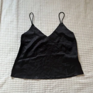 Svart linnetopp från The Black Label - Svart linnetopp i slätt, glansigt material från Lager 157 (The Black Label Collection). Toppen har smala axelband och v-ringad halsringning, vilket ger en stilren och enkel look. Perfekt att styla med jeans eller kjol för en trendig vibe.