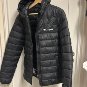 Svart pufferjacka från Champion - Svart pufferjacka från Champion med huva och dragkedja framtill. Jackan har quiltad design och är tillverkad i glansig polyester. Perfekt för kyliga dagar och har en clean look med diskret logga på bröstet.