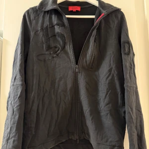 Svart bomberjacka från Hugo Boss - Svart bomberjacka från Hugo Boss med broderade detaljer på bröstet, ärmen och ryggen. Jackan har dragkedja framtill, hög krage och justerbar dragsko nertill. Tillverkad i bomullsmaterial och har en cool streetstil.