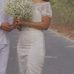 Vit spetsklänning med slits från white one Barcelona - Elegant vit långklänning från white one Barcelona med spets upptill, korta ärmar och vacker slits framtill. Klänningen har figurnära passform, satinliknande kjol och ett bälte med rosett i midjan. Spetsen är dekorerad med diskreta paljetter och knappar i ryggen.