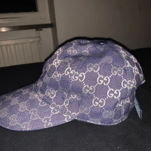 Blå keps från Gucci med GG-mönster - Säljer en blå keps från Gucci med det klassiska GG-monogrammet över hela kepsen. Kepsen har böjd skärm och är tillverkad i ett slitstarkt tyg med tydlig logga. Perfekt för dig som vill sticka ut med en lyxig accessoar.