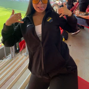 Svart AIK hoodie från Nike - Svart hoodie från Nike med AIK-logga på bröstet och vit Nike-swoosh. Hoodien har huva, dragkedja framtill och två fickor. Tillverkad i mjuk bomullsmix, perfekt för att representera AIK på läktaren eller i vardagen.Nypris 700